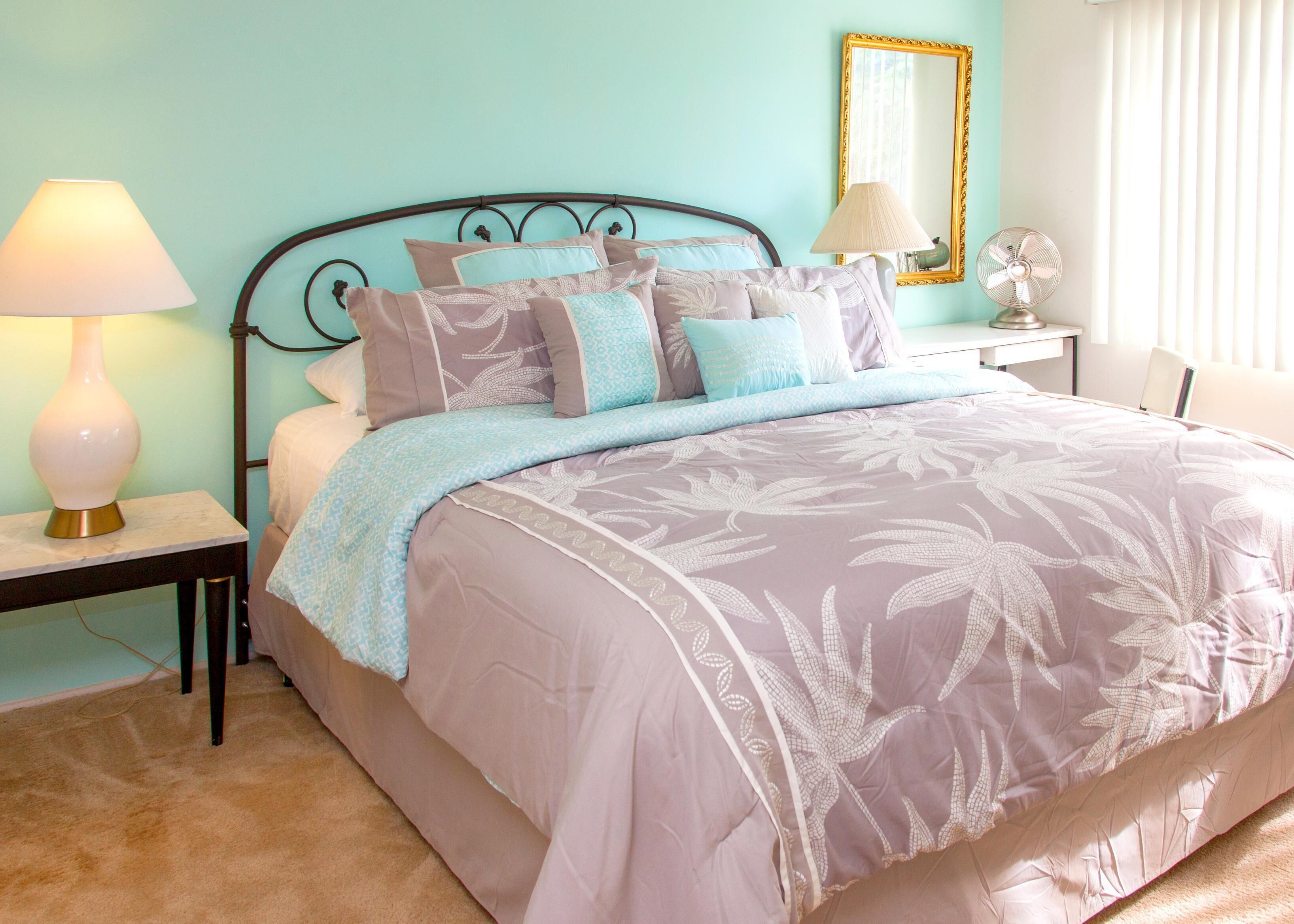 A bedroom with a bed, a nightstand, a lamp, a mirror, and a fan.
