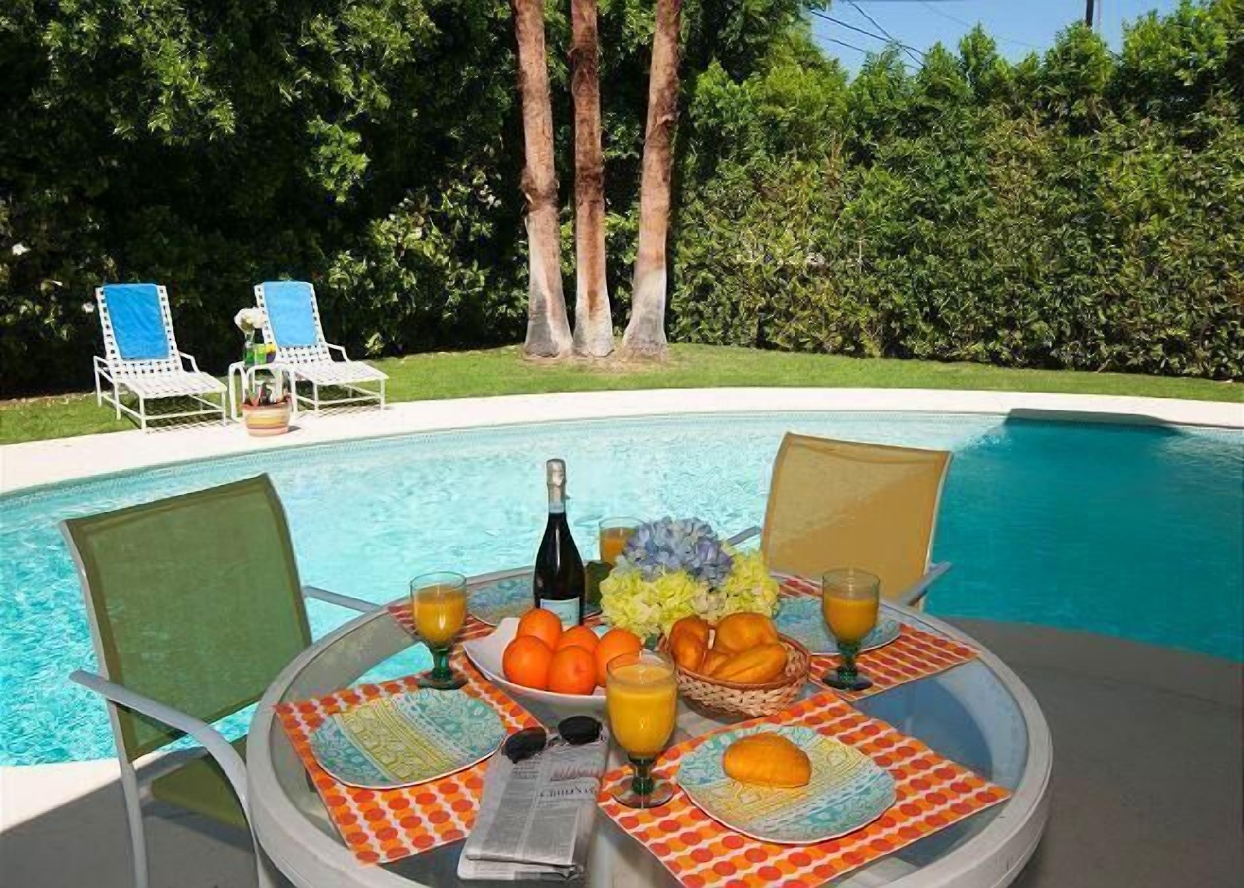Une table placée au bord de la piscine, avec une bouteille de vin, des oranges et des verres de jus d’orange, entourée de chaises longues.