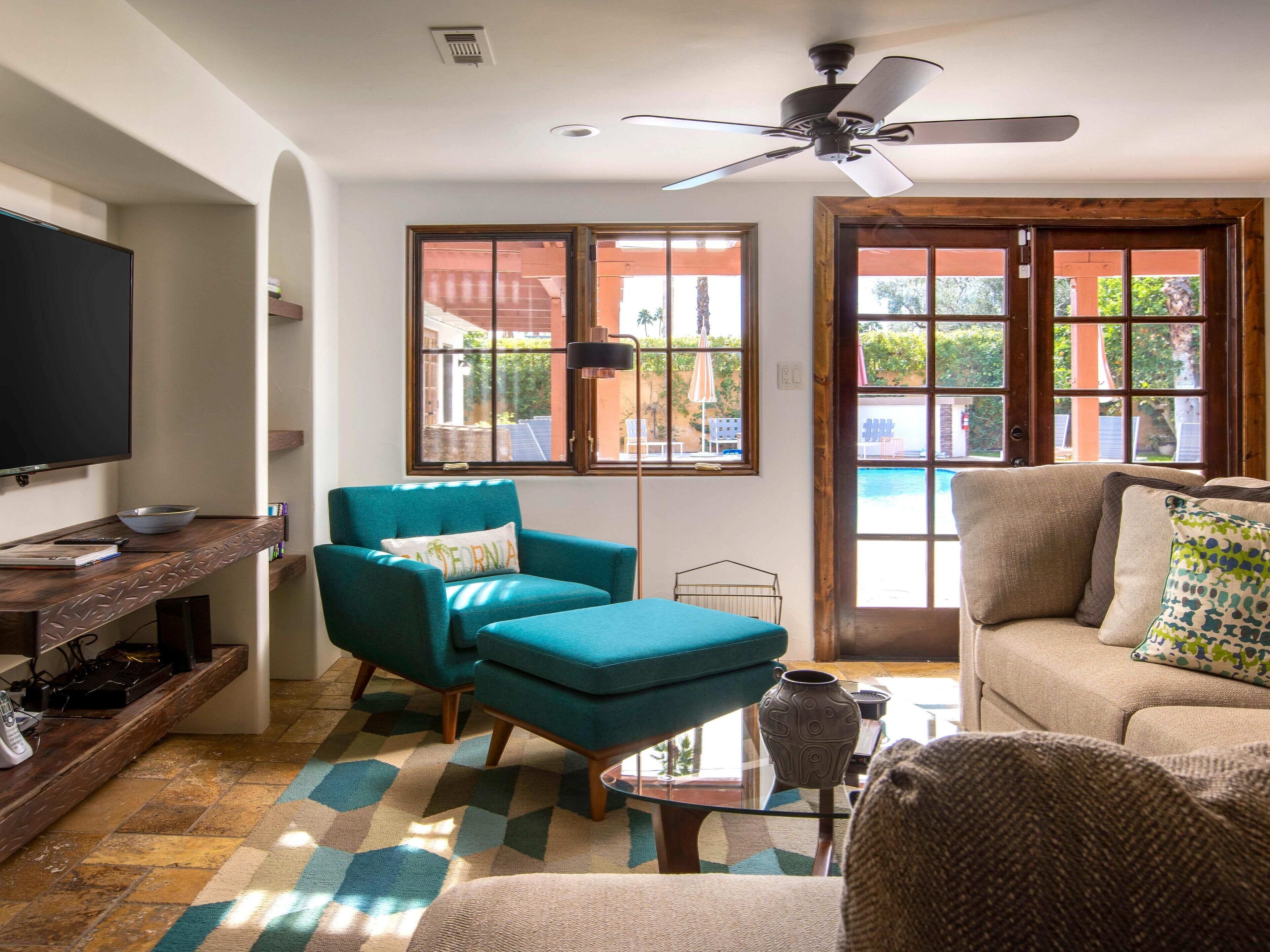 Una sala de estar con sillones color turquesa, un sofá color beige, un televisor de pantalla plana y un ventilador de techo.