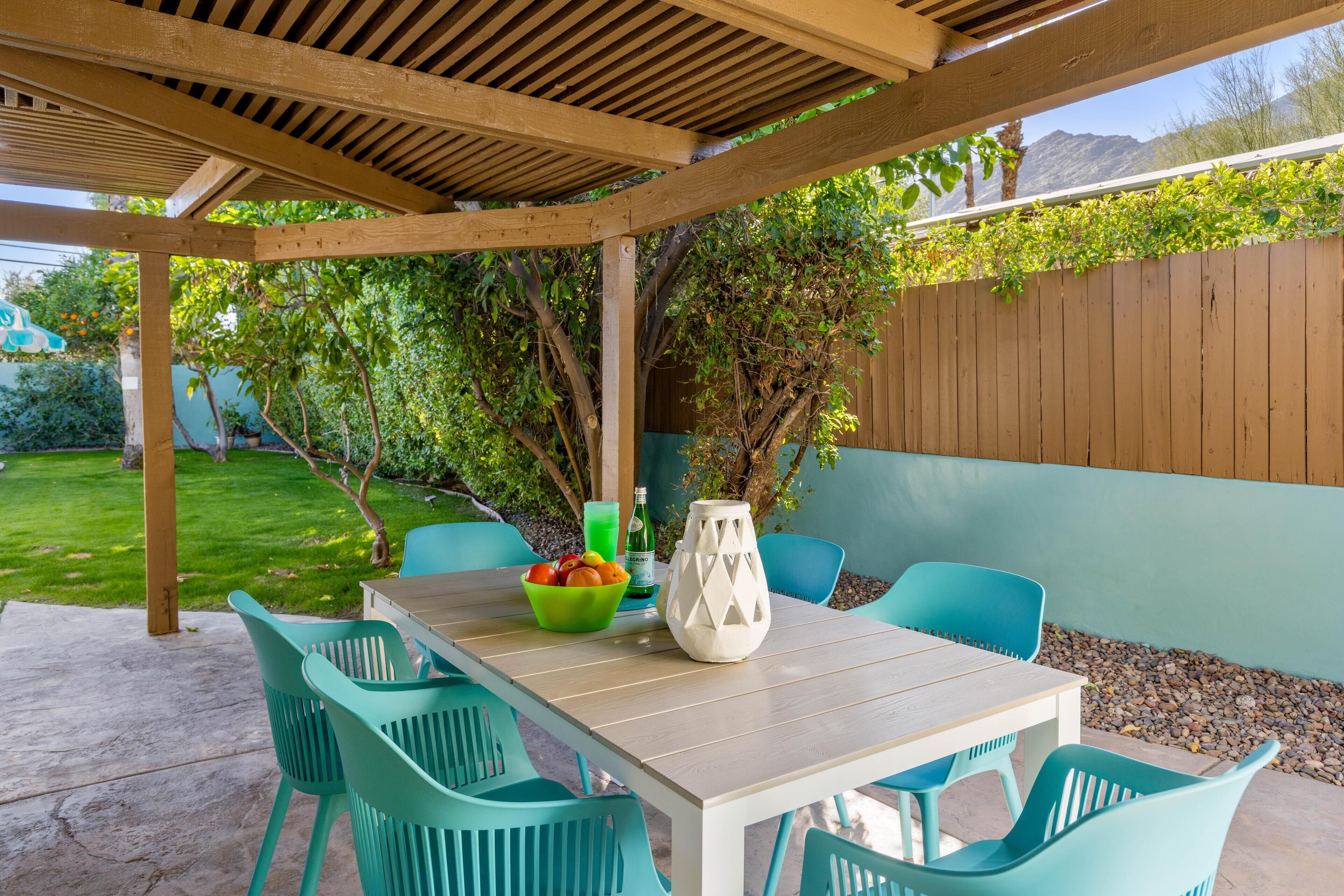 A covered patio with a wooden table and turquoise chairs, surrounded by greenery and a wooden fence.