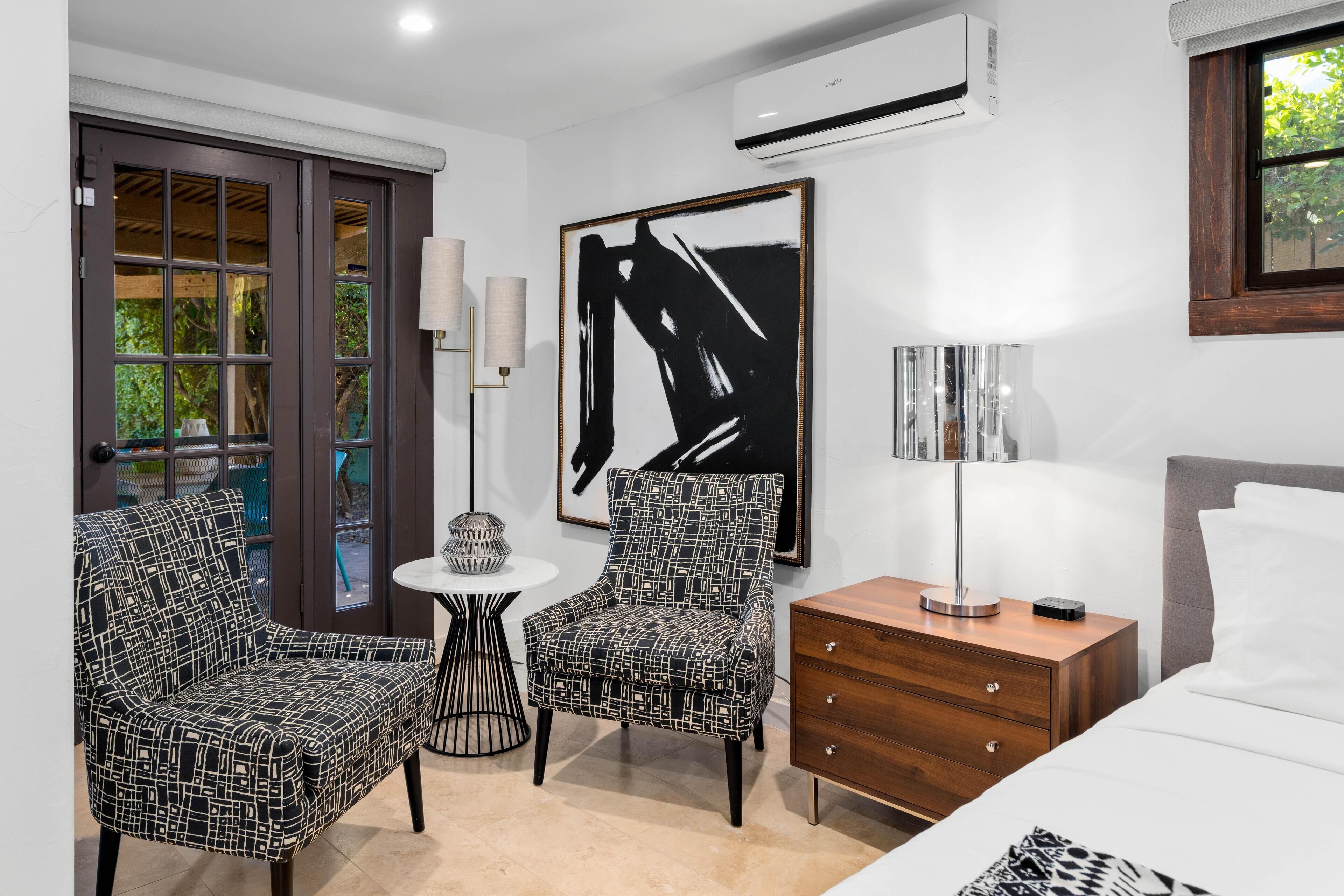 A modern hotel room with a bed, two patterned armchairs, a small table, a lamp, and a large abstract painting.