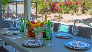 A table set for dining with plates, glasses, and bottles, overlooking a pool and garden with palm trees and pink flowers. - Azure Oasis (Palm Springs)
