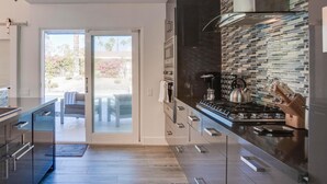 A modern kitchen with a glass door leading to a patio, stainless steel appliances, and a black tiled backsplash. - Azure Oasis (Palm Springs)