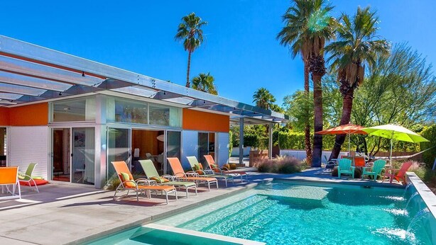 A modern pool area with lounge chairs and umbrellas, surrounded by palm trees and a clear blue sky. - Glass and Steel House 2012 (Palm Springs)