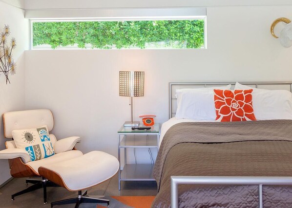 A modern hotel room with a bed, a chair, a nightstand, a lamp, and a window with a view of greenery.