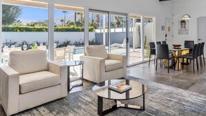 A modern living room with a sofa, coffee table, and dining area with chairs and a table. Large windows offer a view of a pool and greenery. - Happy Hour (Palm Springs)