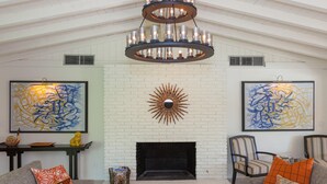 A spacious living room with a white brick fireplace, two large abstract paintings, a modern chandelier, and comfortable seating.
