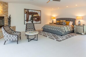 A bedroom with a bed, two nightstands, a chair, a table, a fireplace, and a large painting. - Hidden Haven (Palm Springs)