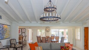 A spacious living area with a grey sectional sofa, a coffee table with a book and a plant, and a chandelier. There is a dining area with chairs and a table, and a large painting on the wall.