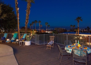 A deck with outdoor seating, a table set for dining, and a view of the city at dusk. - Hilltop Haven (Palm Springs)