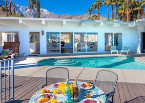 A modern poolside area with a clear view of a mountain range, a pool, and a dining table set with fruits and beverages. - Hilltop Haven (Palm Springs)