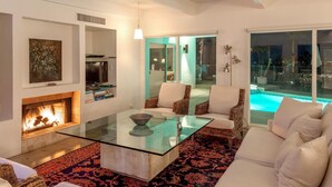 A modern living room with a glass coffee table, white sofas, a fireplace, and a view of a swimming pool at night. - Hilltop Haven (Palm Springs)