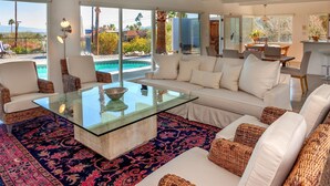 A spacious living room with a glass coffee table, white sofas, and a large window overlooking a pool and outdoor area. - Hilltop Haven (Palm Springs)