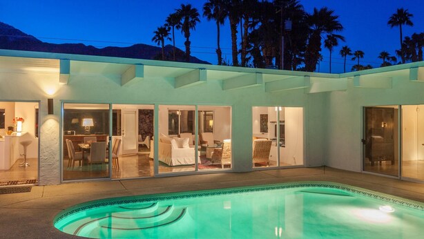 A modern house with a swimming pool, glass doors leading to a dining area, and palm trees in the background. - Hilltop Haven (Palm Springs)