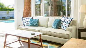 A living room with a sofa, a coffee table, and a lamp. There is a window with a view of a pool and greenery outside. - Canary Palms (Palm Springs)