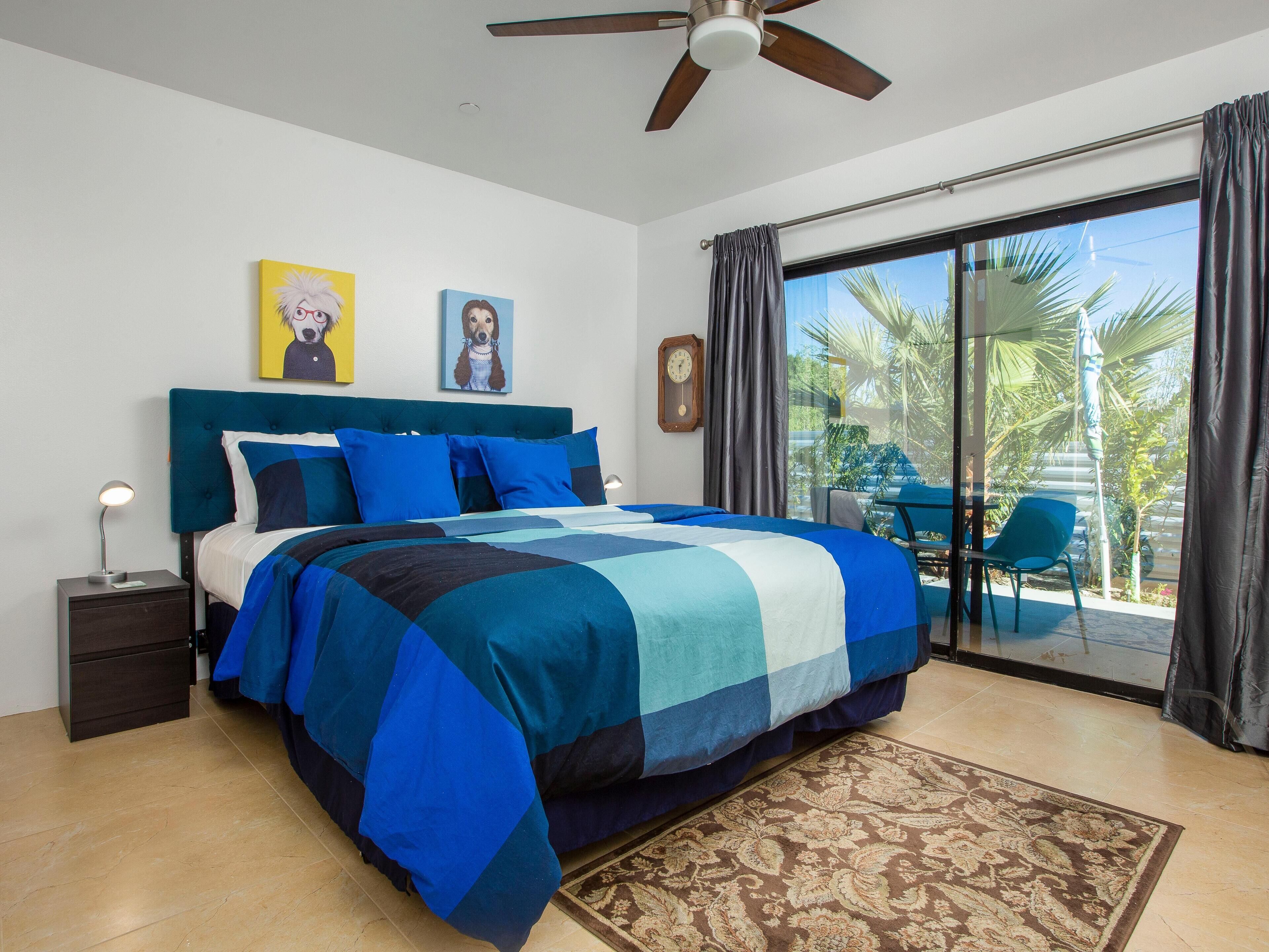 A bedroom with a bed, a nightstand, a ceiling fan, a window with curtains, and a view of the outdoors.