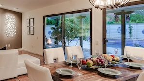A modern dining area with a large table set for six, surrounded by white chairs, and a chandelier above. - Andiamo al Sole (Palm Springs)