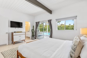 A modern bedroom with a large bed, a flat-screen TV, a dresser, and a view of a pool through a sliding glass door. - Alexander on Bedford (Palm Springs)