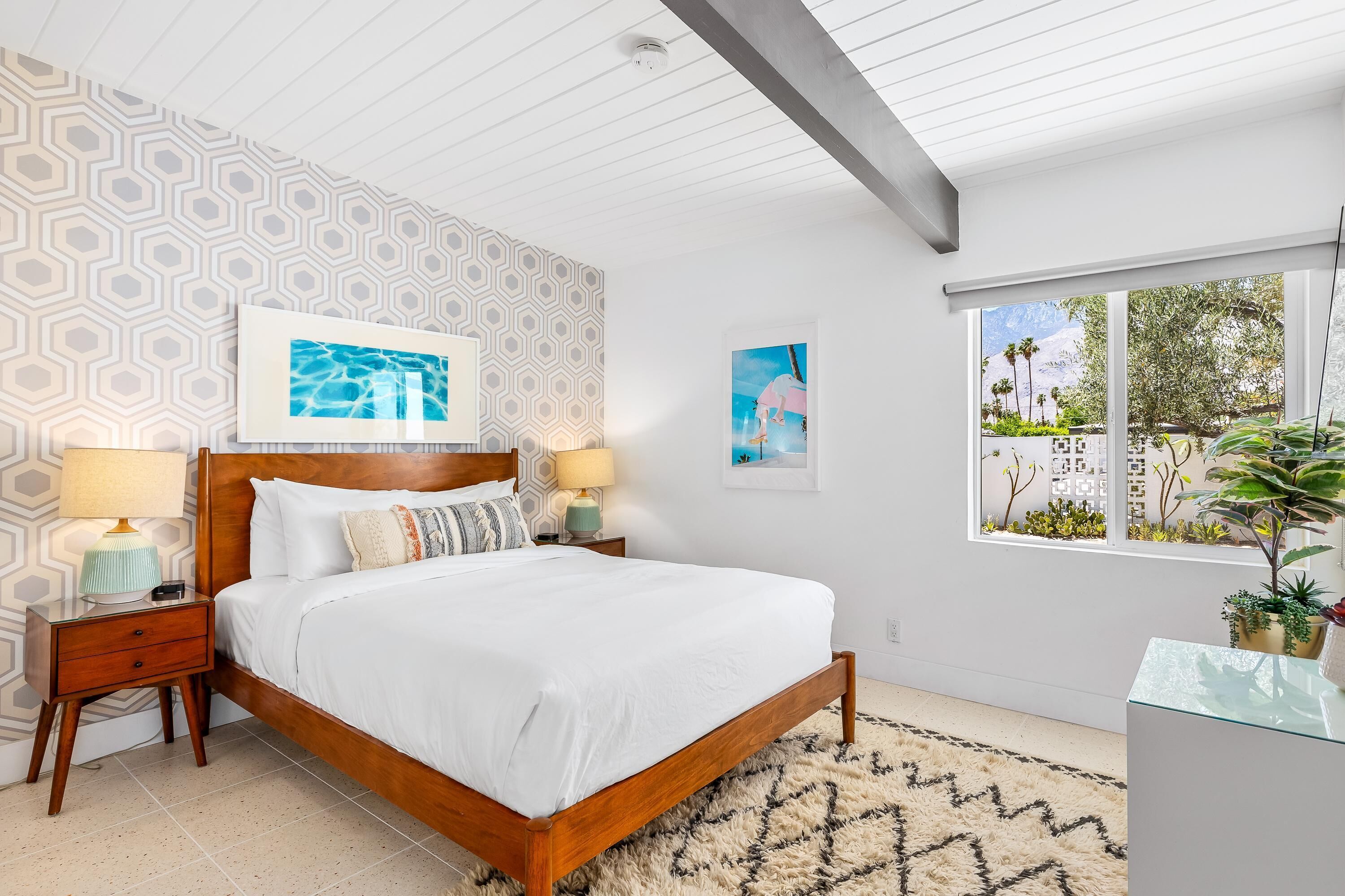 A bedroom with a wooden bed, two bedside tables with lamps, a patterned wall, and a window with a view of greenery and buildings.