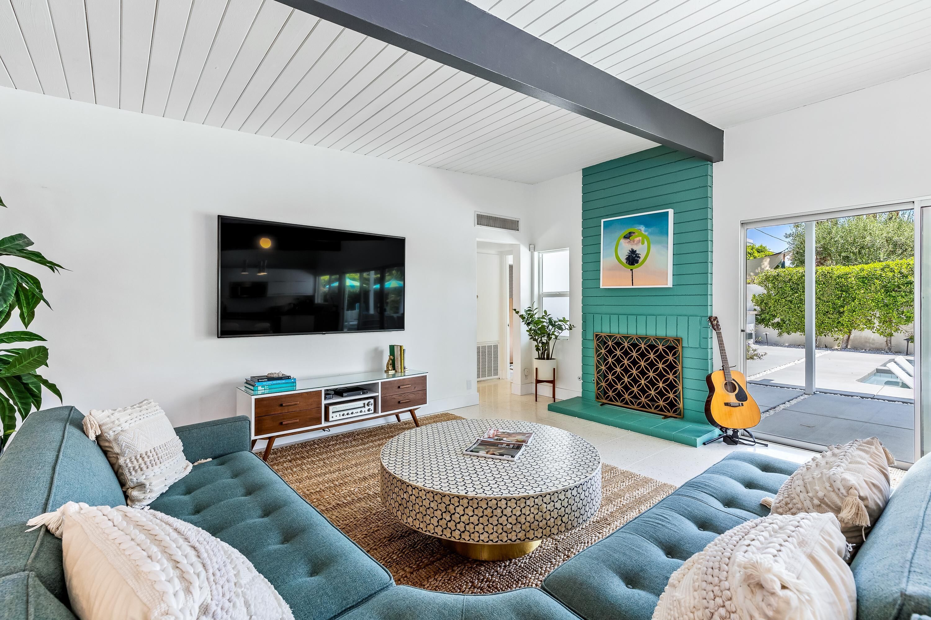 Un salon moderne avec un grand canapé d’angle, une table basse ronde, un téléviseur à écran plat et une guitare appuyée contre un mur vert.