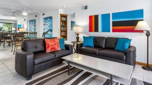 A modern living room with two black leather sofas, a white coffee table, and a striped rug. There are bookshelves, a dining area, and artwork on the walls.