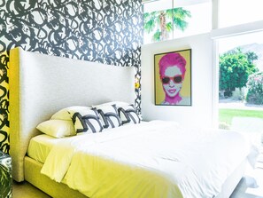 A bedroom with a bed, pillows, a headboard, a wall with a patterned wallpaper, and a window with a view of greenery. - PS Smile (Palm Springs)