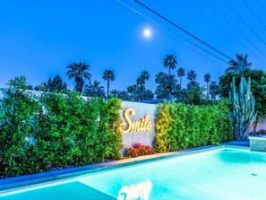 A swimming pool with a neon 'Smile' sign, surrounded by lush greenery and palm trees. - PS Smile (Palm Springs)