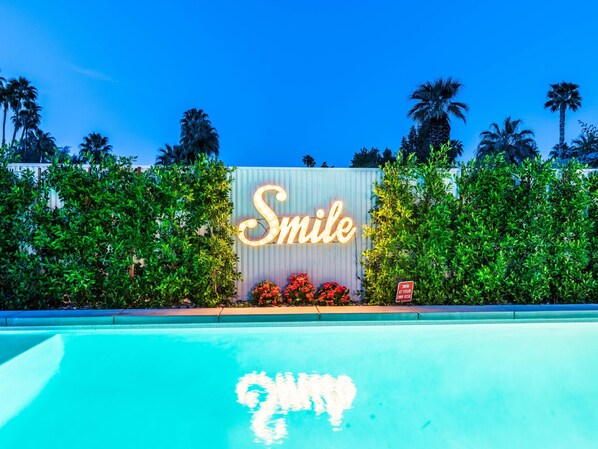 A swimming pool with a neon 'Smile' sign, surrounded by lush greenery and palm trees. - PS Smile (Palm Springs)