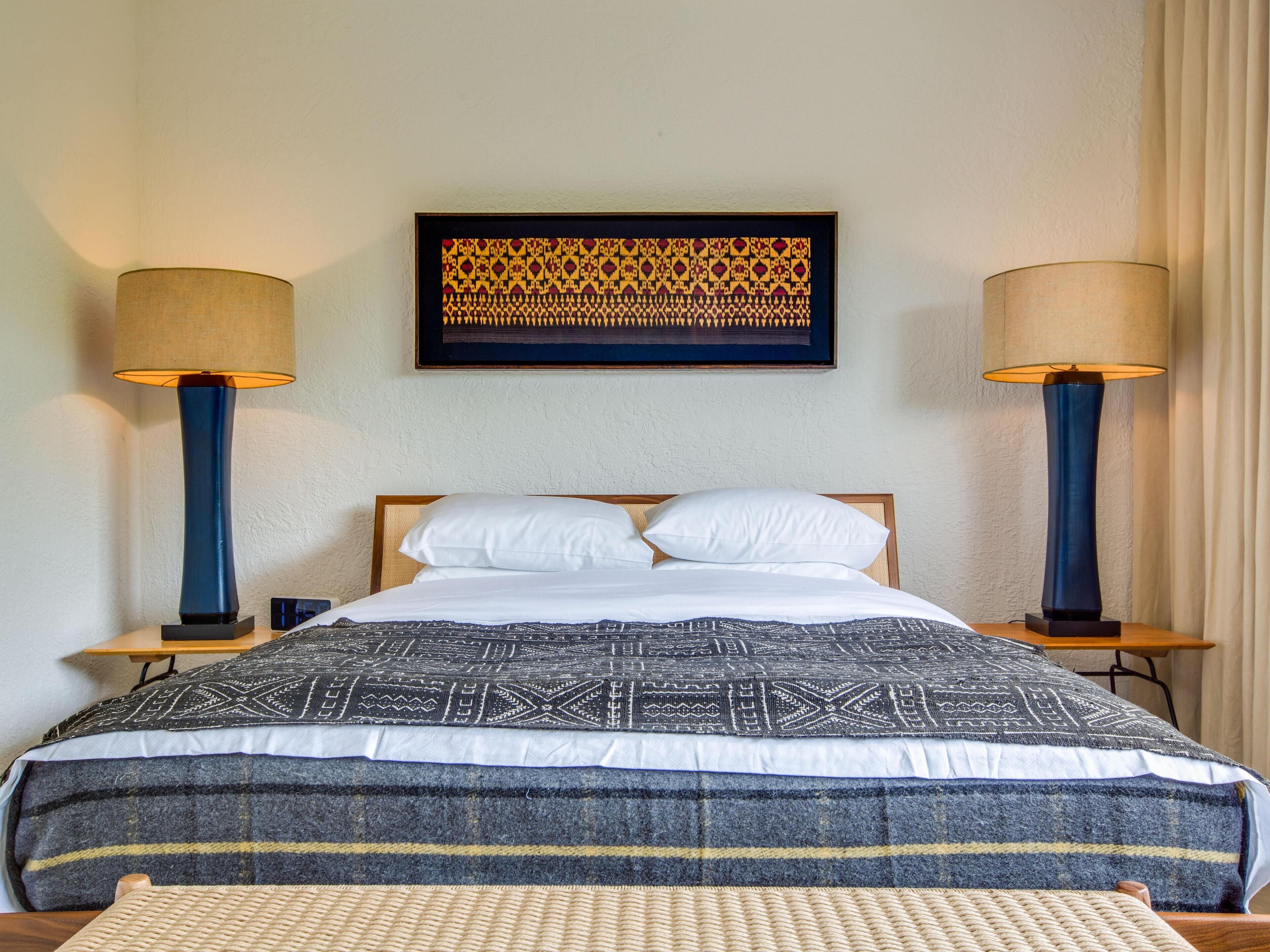 Een netjes opgemaakt bed met een bedsprei met patroon, met aan weerszijden nachtlampen en een lijstwerk erboven.