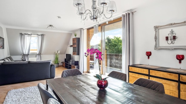 A modern living room with a dark wooden dining table, a black sofa, and a large window with curtains.
