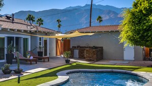 A backyard with a swimming pool, a patio with a table and chairs, a stone barbecue area, and a view of mountains. - Boho Rhapsody (Palm Springs)