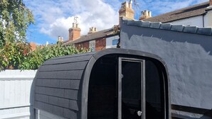 A black, curved structure with a glass door, likely a shed or storage unit, in a residential backyard with a white fence, green lawn, and brick houses in the background. - The Railway Inn MK | Sauna & Jacuzzi | Family & Pet-Friendly | Sleeps upto 8 (Milton Keynes)