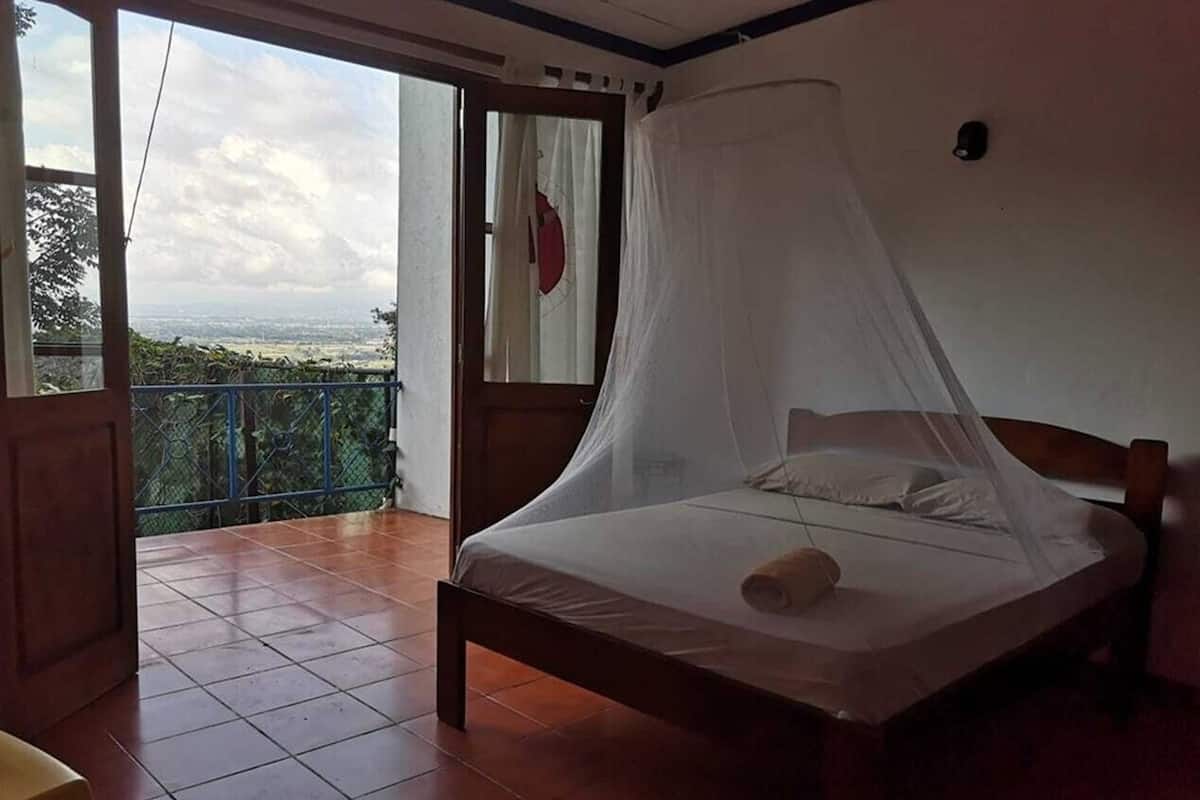 A bedroom with a bed under a mosquito net, a balcony with a view, and a door with a curtain.