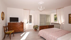 Ein Schlafzimmer mit einem Holzkasten, einem Bett mit gemustertem Bettbezug, einem Fernseher auf dem Kasten, einer Topfpflanze und einem Fenster mit Vorhängen.