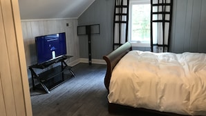 A bedroom with a bed, a TV on a stand, a window with shutters, and wooden flooring.