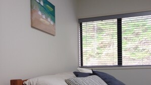 Una camera da letto con un letto, un comodino in legno, una cassettiera, una finestra con tende e un quadro appeso al muro.