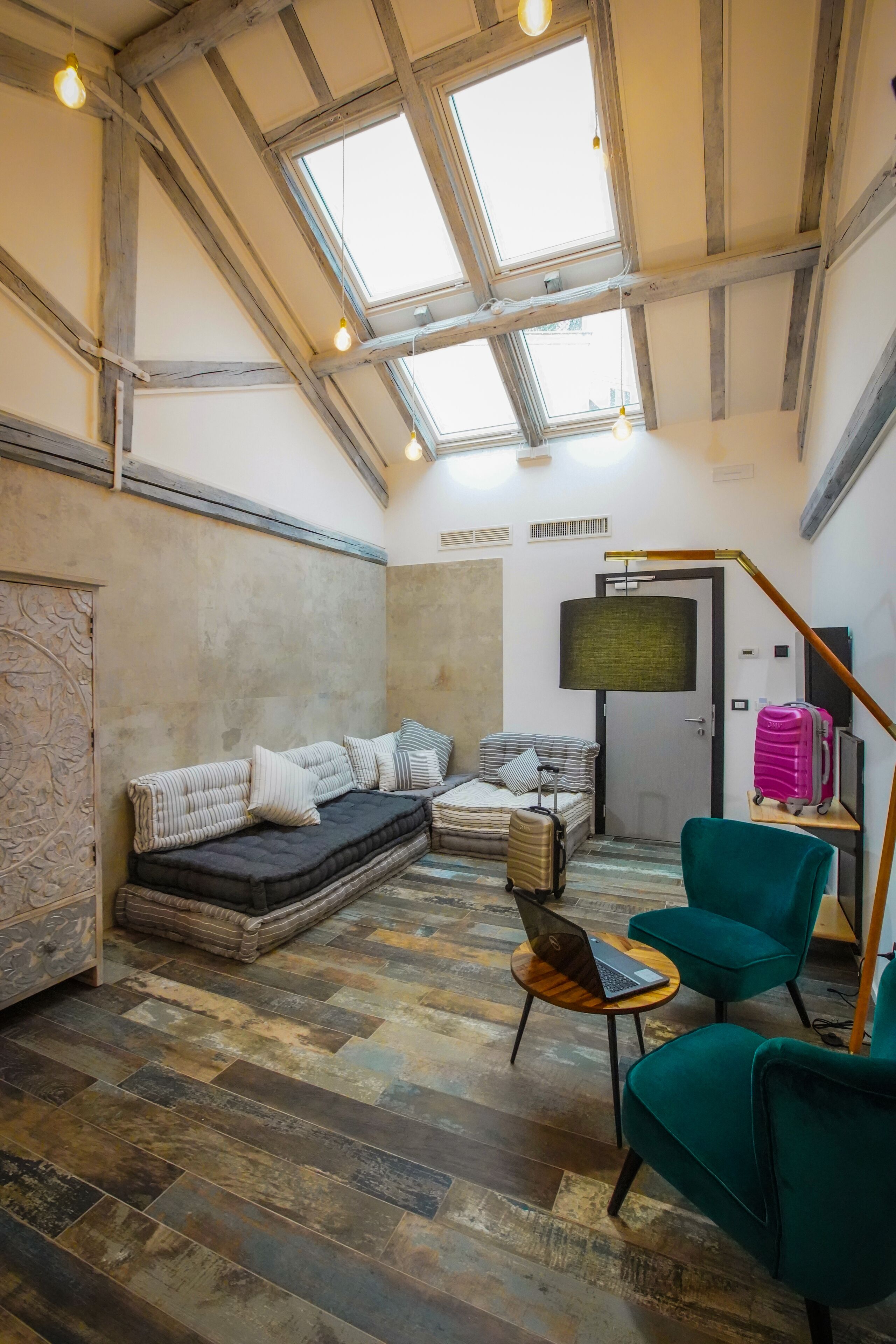 Habitación estilo loft con techo alto, vigas a la vista, tragaluz grande, sofá modular, sillón verde, mesa redonda pequeña con una computadora portátil y una maleta.
