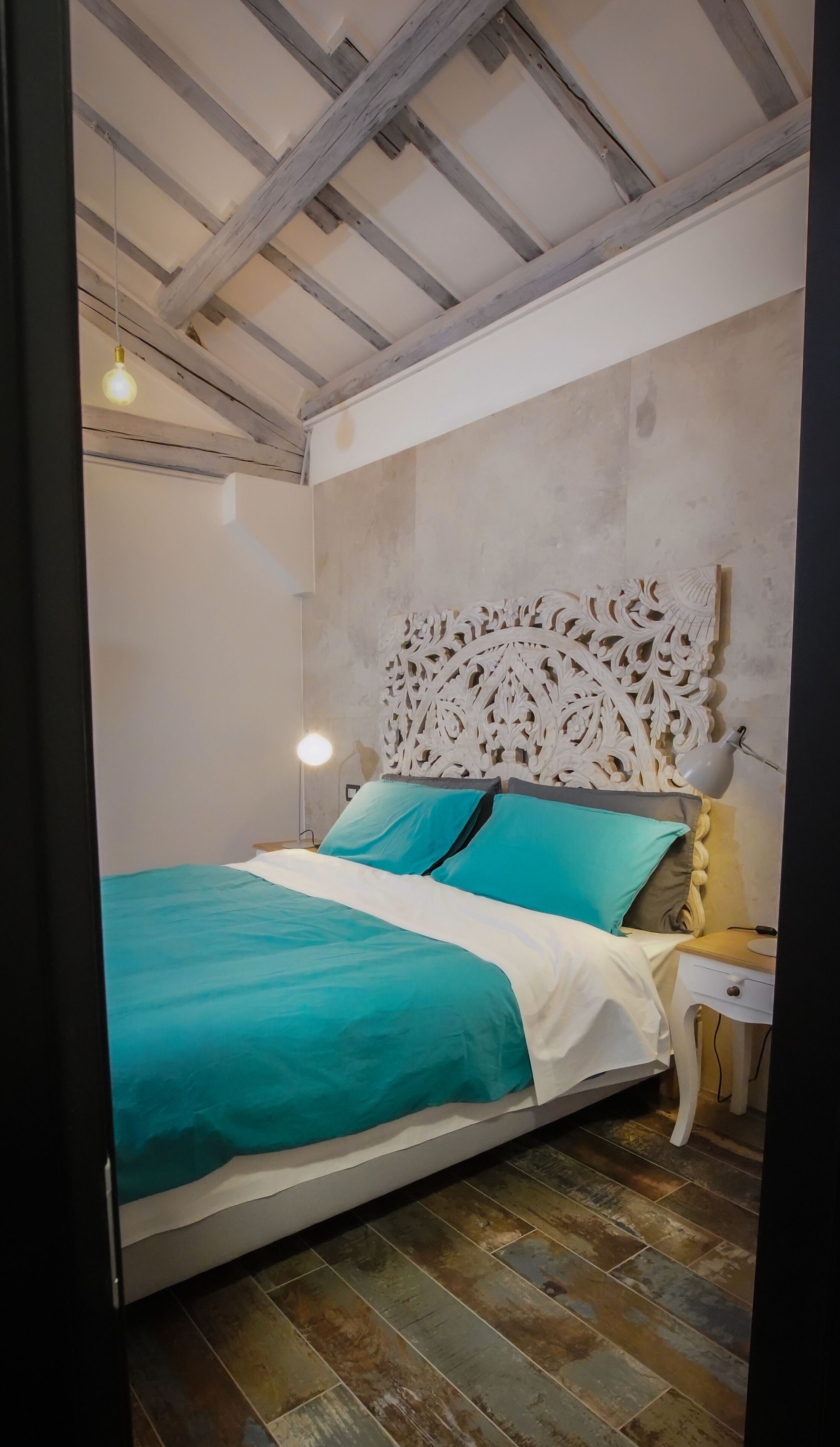 Un dormitorio con una cama, una mesita de noche, una lámpara de pared y un techo con vigas a la vista.
