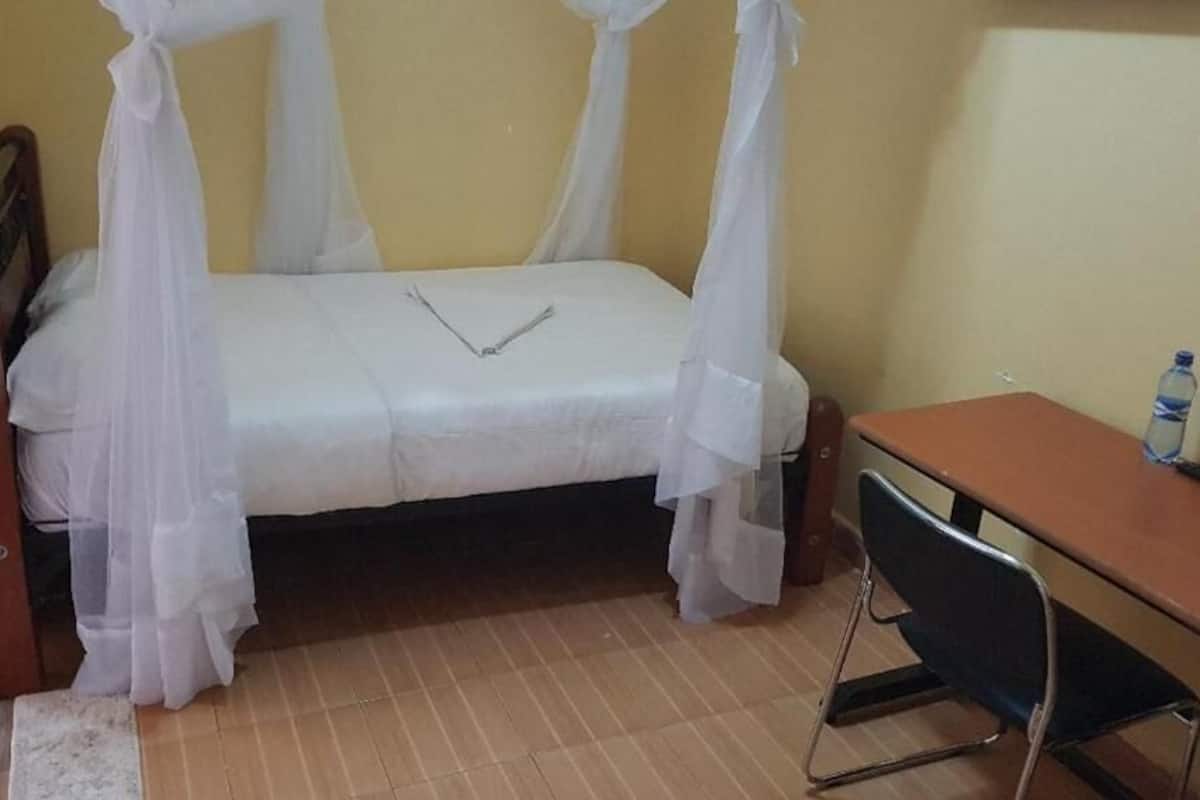 Uma cama de solteiro com dossel, uma escrivaninha de madeira, uma cadeira e uma garrafa de água sobre a escrivaninha.