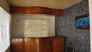 A reception area with a wooden reception desk, a clock, a window with metal bars, and a television displaying a news program. - Lila Hotel (Kisumu)