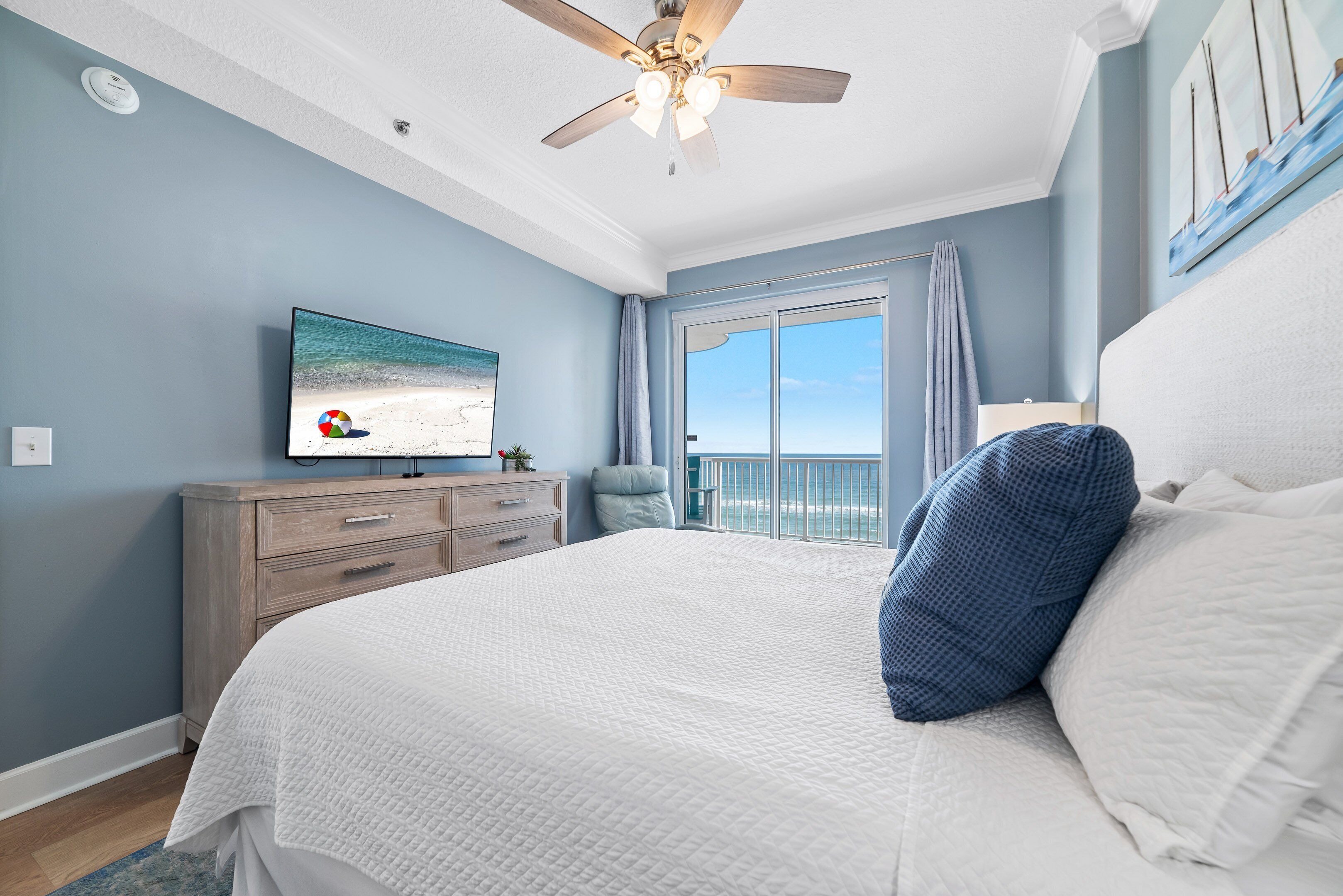 A bedroom with a large bed, a ceiling fan, a TV on the wall, and a view of the ocean through the window.
