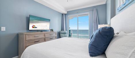 A bedroom with a large bed, a ceiling fan, a TV on the wall, and a view of the ocean through the window.