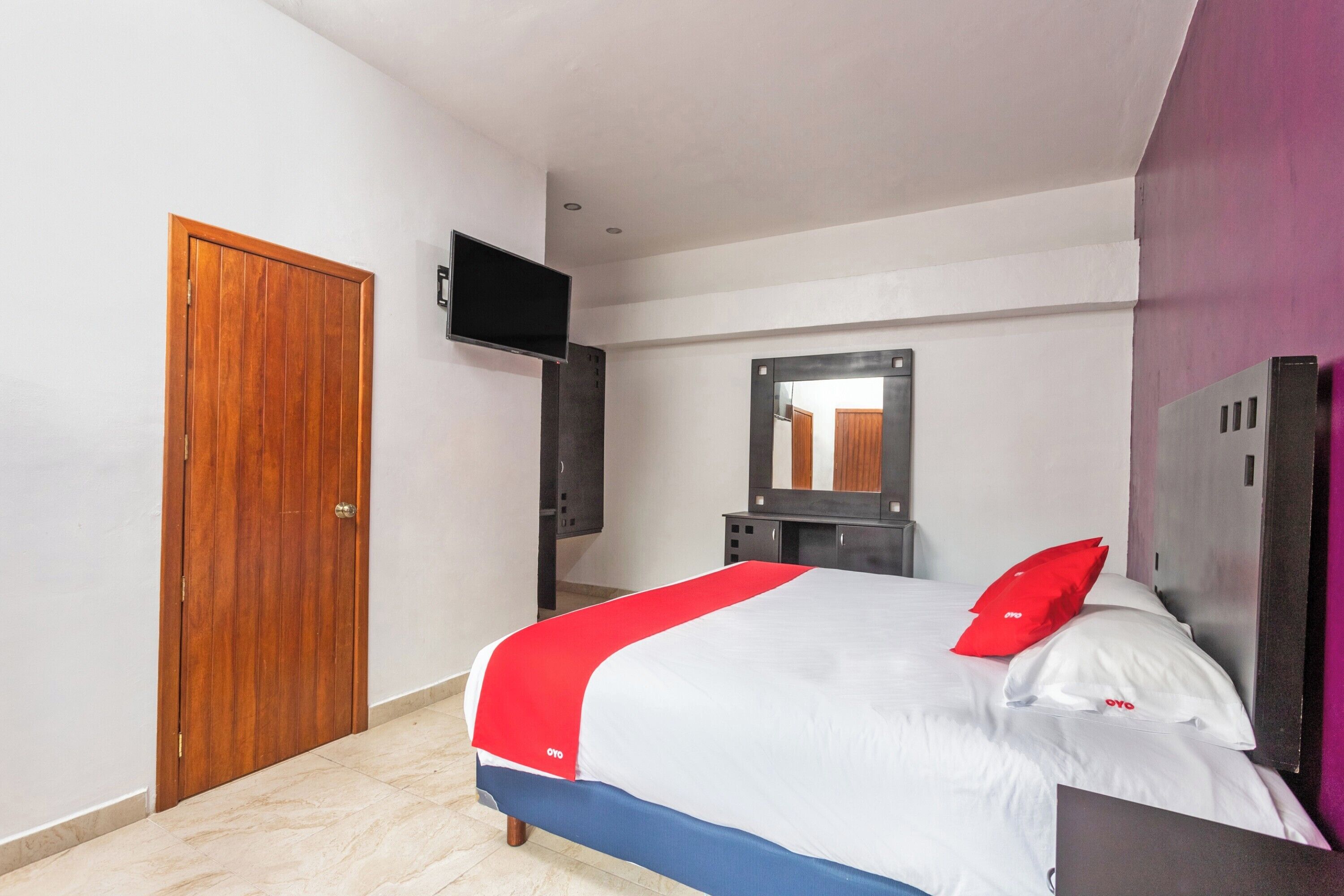 Un dormitorio con puerta de madera, cama con ropa de cama blanca y roja, televisor montado en la pared y un espejo sobre la cama.