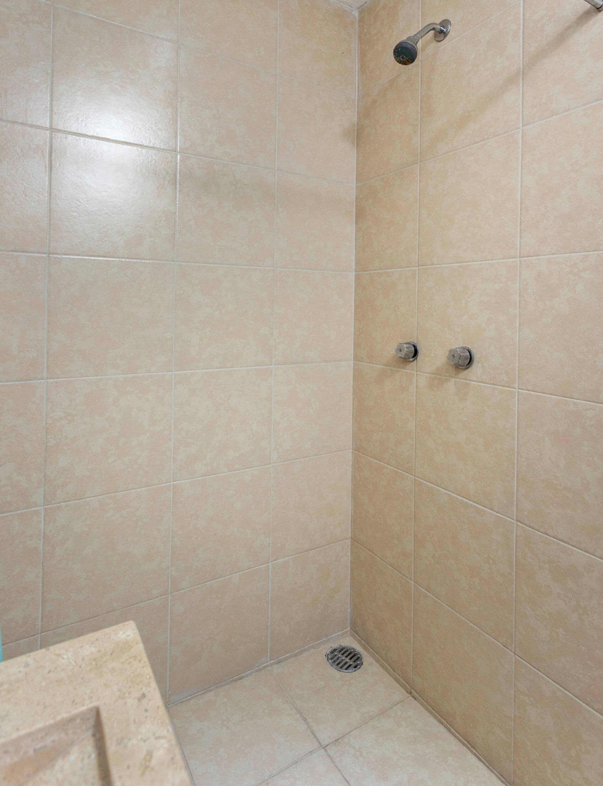 A bathroom with beige tiles, a shower with a handheld showerhead, and a drain.