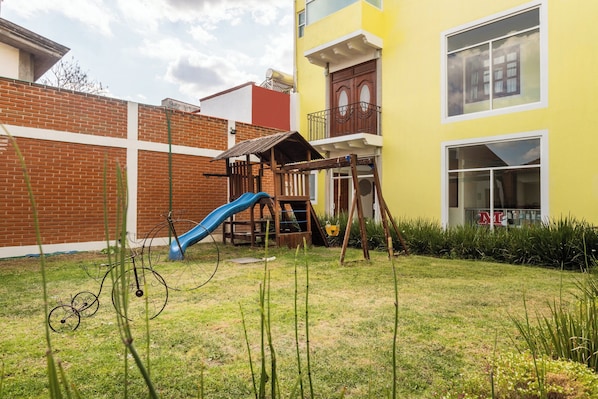 Zona residencial con un muro de ladrillo, un parque infantil de madera y un edificio amarillo con balcón.
