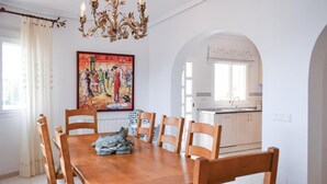 Ein Essbereich mit Holztisch und -stühlen, einer Kronleuchter, einem Gemälde an der Wand und einer durch einen Bogen sichtbaren Küche.