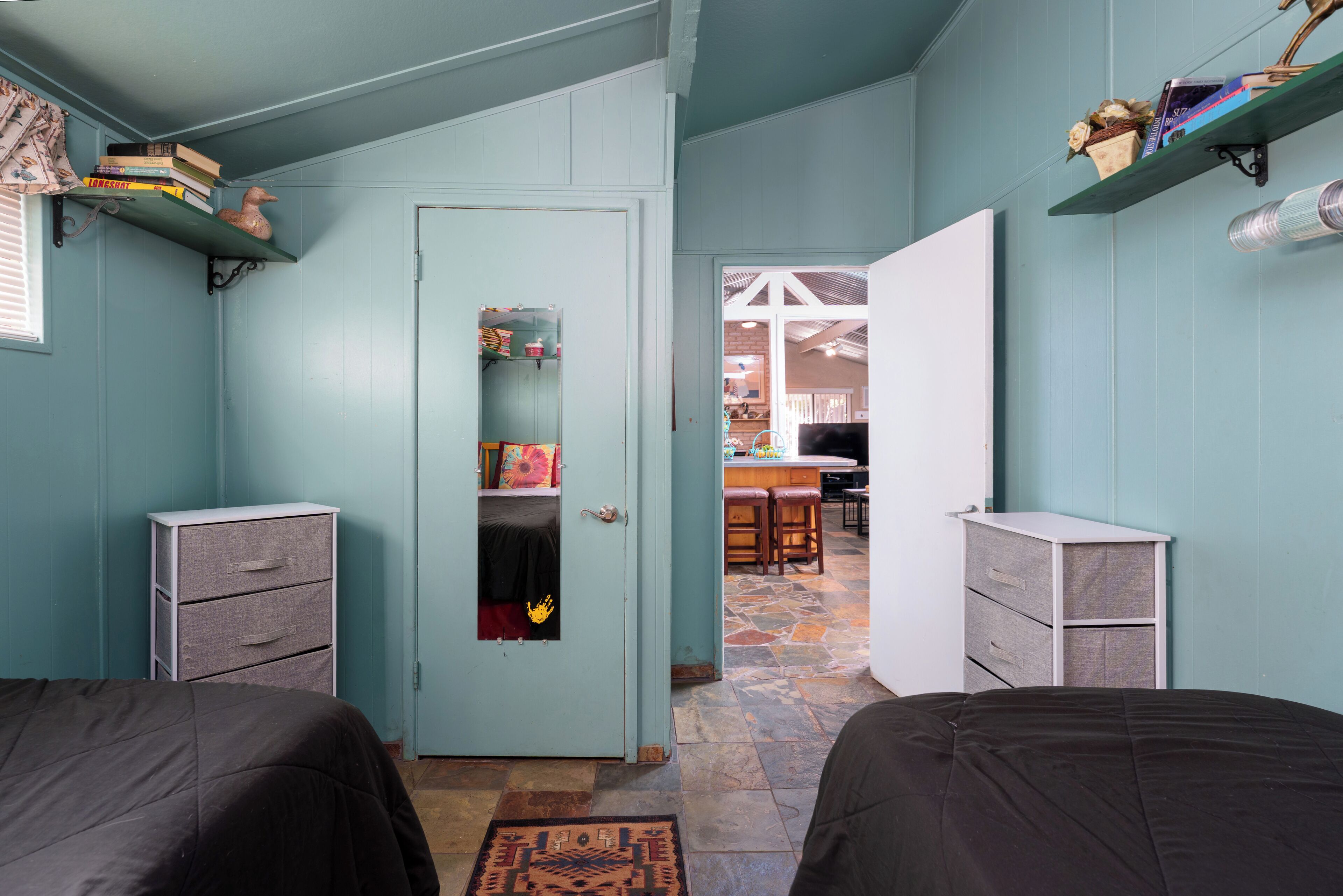 A room with teal walls, two beds, a door with a mirror, and a kitchen area visible through an open door.