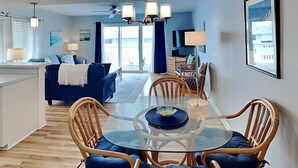 Un salon moderne avec une table à manger en verre, des chaises rembourrées en tissu bleu, un canapé et un téléviseur.