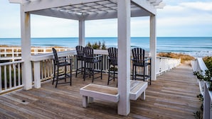 Una terraza de madera con pérgola, sillas y un banco, con vista al océano.