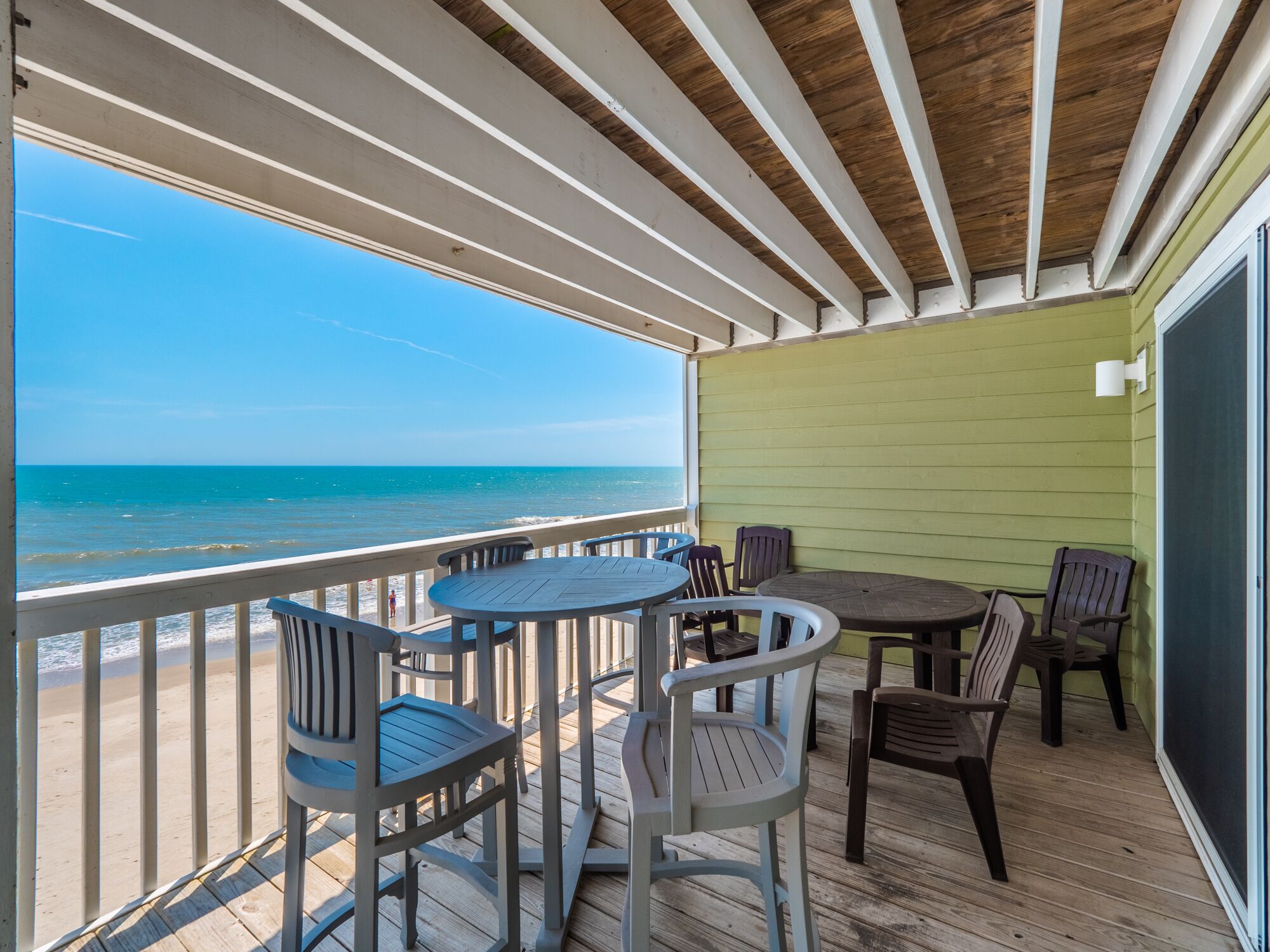 Een overdekt buitengebied met uitzicht op het strand, met een houten terras, verschillende tafels en stoelen, en een groene wand.